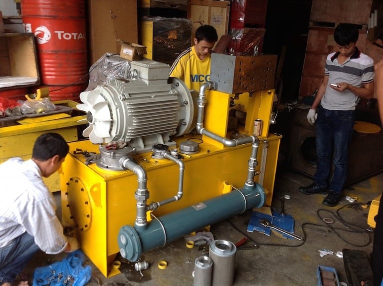 Technicians assembling a large yellow hydraulic pump system in an industrial workshop with electrical motor and piping components