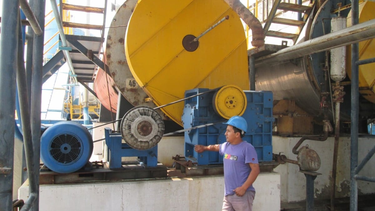 Worker in purple shirt standing near large industrial machinery with yellow pulley and blue motors in factory setting