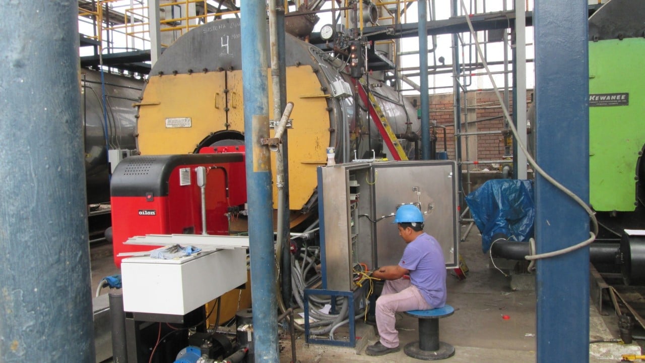 Worker in blue hard hat servicing industrial machinery including yellow and red equipment in a factory workshop.