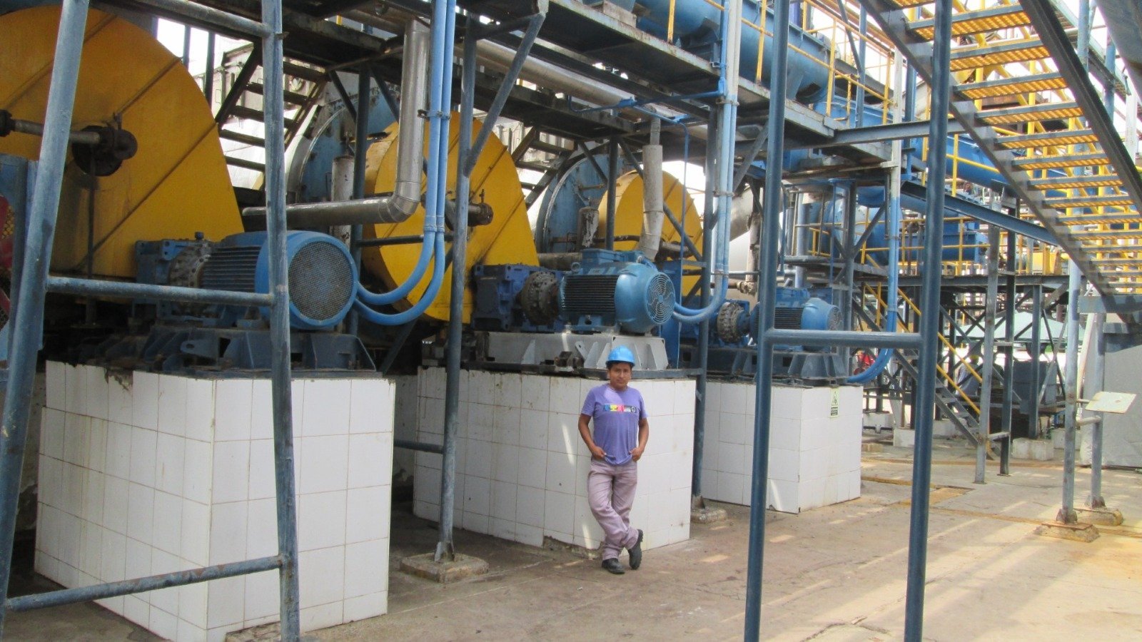 Worker standing beside large yellow industrial machinery in a factory with white protective barriers and steel framework.