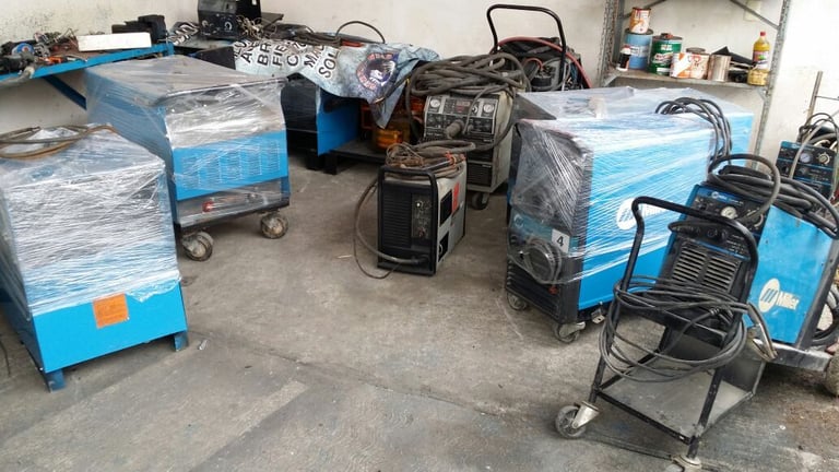 Industrial welding equipment and machinery stored in a workshop, including blue and black welders wrapped in plastic.